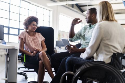 Businessman discussing with female colleagues in office. Disabled businesswoman is sitting in wheelchair with executives at brightly lit workplace. They are in smart casuals.