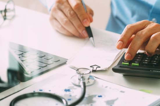 Image de mains masculines. Dans une main, il tient un stylo sur une pile de papier, une calculatrice d’un côté et un ordinateur de l’autre. Son stéthoscope est assis devant