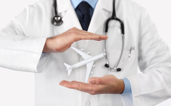 Torso of a male doctor in a white lab coat. He is wearing a stethoscope around his neck and cradling a model airplane between his hands. The image supports the concept of out-of-country medical assistance.