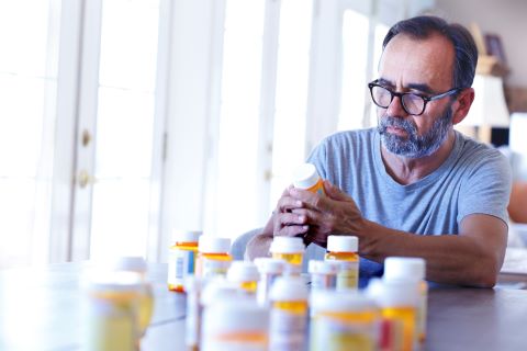 A latino baby boomer sits at his dining room table and sorts through various prescription medications as sunlight filters in through the window behind him bathing the room with a soft glow of light.