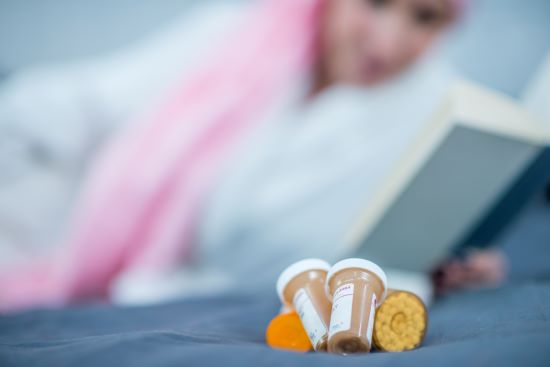 A Caucasian woman is reading in bed. She is wearing a head scarf to hide hair loss from chemotherapy. Pill bottles are in the foreground.