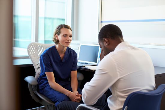 Female Caucasian nurse in consultation with a depressed African American man who sits with his head in his hands. 