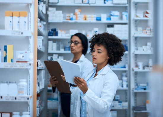 Conduire une pharmacie performante avec un travail d'équipe - Stock photo d'une jeune femme inventoriant une pharmacie sur une tablette numérique avec son collègue à l'arrière-plan.