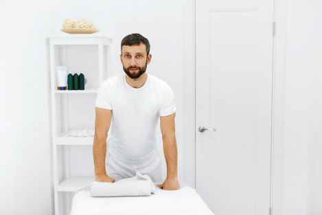 Caucasian male massage therapist standing and leaning forward with his hands resting on the massage table.