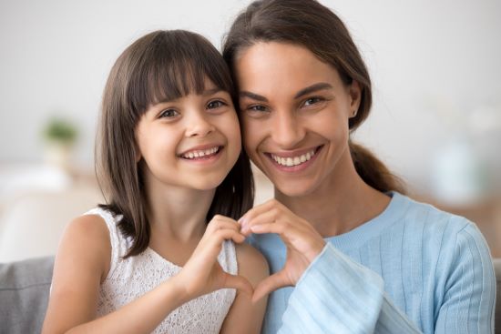 Cute little daughter and happy mother join hands in shape of heart as concept of mom and child love care support, smiling mum and her kid girl looking at camera posing together for headshot portrait - used to represent healthy and happy, and great smiles thanks to good dentistry.