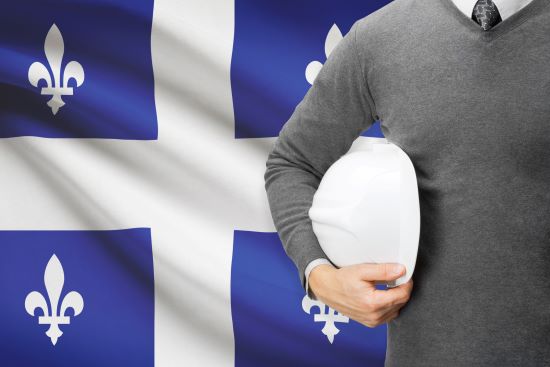 Torso of a Caucasian man holding a white hard hat standing in front of a Quebec flag.