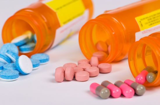 Multi-coloured pills spilling out of orange pill bottles onto a white table. There are blue and white tablets, pink tablets and pink and brown capsules.