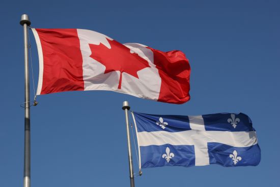 The Canadian and Quebec flags flapping in a breeze against the backdrop of a deep blue sky.