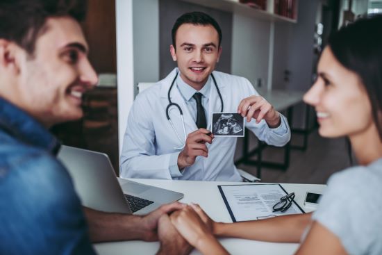 Jeune couple chez le médecin. Ils se tiennent par la main et se sourient tandis que le médecin présente l'image ultrasonde de leur futur bébé conçu grâce à des traitements de fertilité couverts par leur régime d'assurance collective.