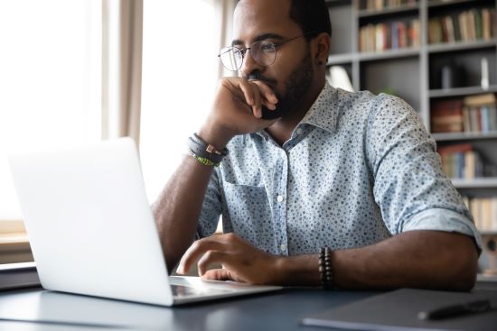 Thoughtful millennial biracial man in eyeglasses staring at laptop screen contemplating a difficult decision.