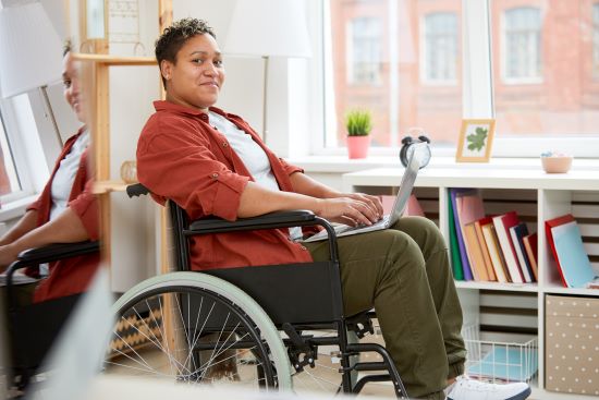 A young black woman is sitting in a wheelchair working on a laptop in a workplace.