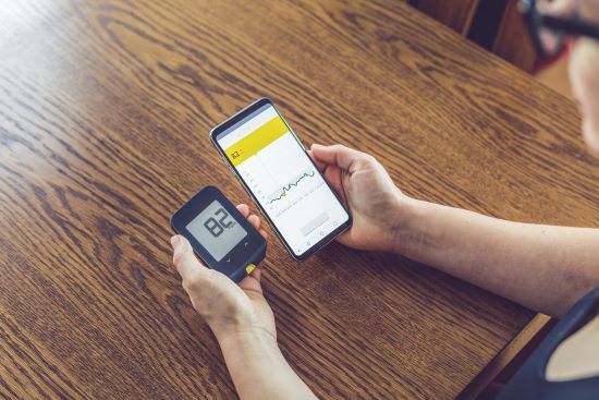 Hands holding a glucose monitor and a cellphone showing blood sugar tracking. Background is a wooden table.