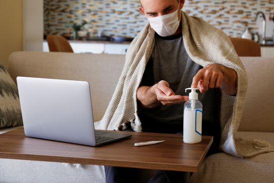 A caucasian male sitting on his sofa with a blanket draped over his shoulders. He is wearing a mask and cleaning his hands with sanitizer. A laptop and digital thermometer sit on the table in front of him.