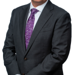 Portrait photo of Eckler Principal August Cruickshanks. He is standing with is hands clasped in front of him looking directly into the camera wearing a black suit and a dark purple tie.
