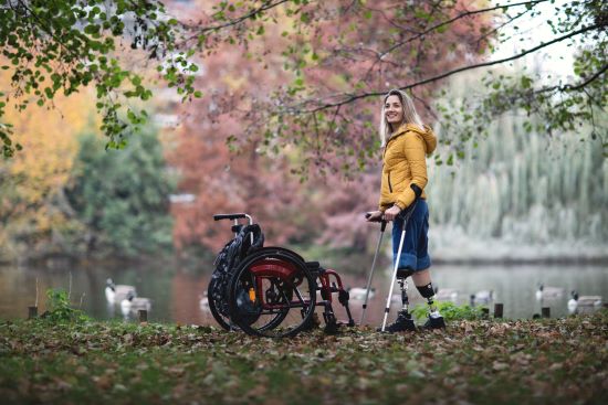 A young girl with prosthetic legs standing next to her wheelchair with the aid of crutches enjoying a nice fall day next to a pond.
