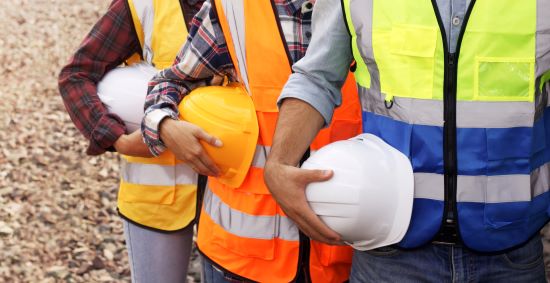 Three constructions workers lined up one in front of the other. The pics shows only the torsos of the workers. They are wearing safety vests and holding hard hats. 