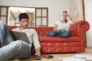Un couple assis sur un canapé en cuir rouge, l'un utilisant un ordinateur portable pour calculer son budget et l'autre regardant ses factures, dans un salon confortable avec des planchers en bois et un grand miroir.