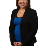 Photo of Eckler Principal Kerry Abehera. She is wearing a black pant suite with a royal blue top and is smiling whiling looking straight into the camera. She has her hands clasped together.
