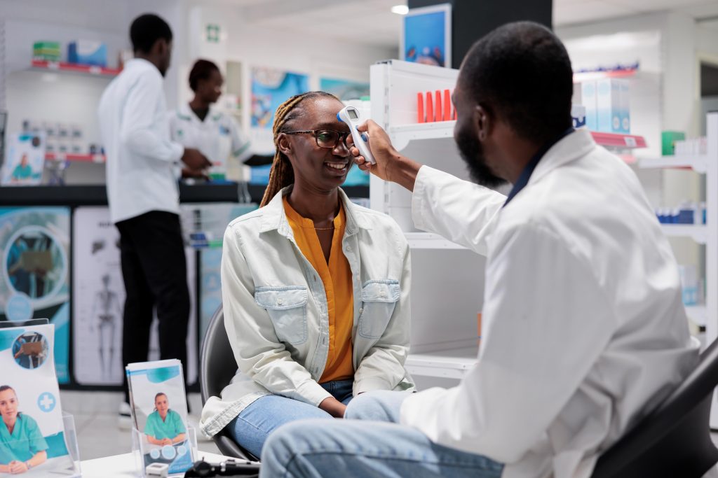 A young black women is undergoing a basic medical examination, conducted by a black male pharmacist, in a pharmacy. The pharmacists is holding an infrared thermometer to her forehead. 