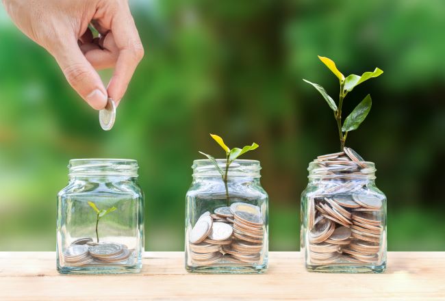 Pictured are three jars against a blurred background of trees and in the jars are coins to represent savings. The first jar has a few coins, the second has more coins and a small tree growing out of them. The third jar is filled almost to the top with coins and the tree has grown substantially as well. 