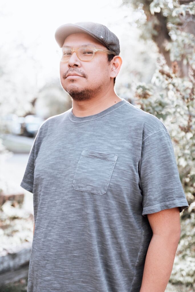 Indigenous artist Torrie Ironstar outdoors among trees and greenery, dressed casually in a gray short-sleeve T-shirt with chest pocket and a flat cap.